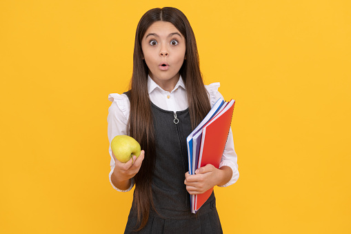 Surprised girl child hold apple and school textbooks yellow background, homework.