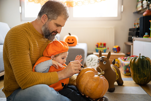 Cute Caucasian toddler boy dressed as a pumpkin, for a Halloween, drawing on pumpkin with his father