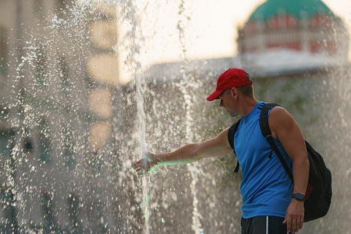 Handsome man dry fountains in the hot summer day in the city Moscow. Time to cold. Fresh water splashes, He touching water sprays. Concept of the leisure, freshness and happiness. Side view