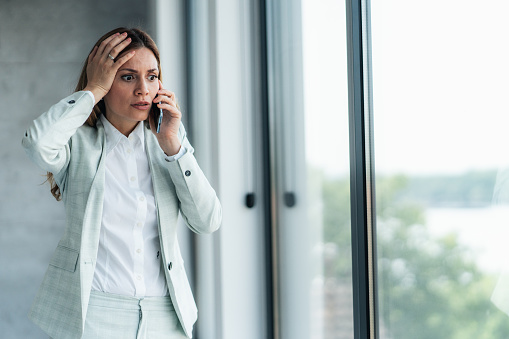 Shot of a young businesswoman looking stressed while talking on the phone. Scared shocked woman talking on phone, receiving bad news, having bad unpleasant conversation with coworker or client.