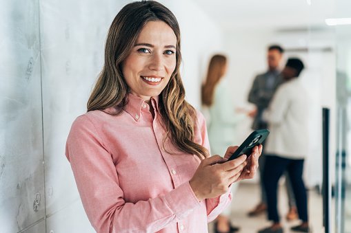 Young beautiful business woman texting in the office. Smiling woman with smart casual businesswear using smartphone for business communication on surfing the net while take a break
