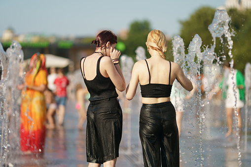 Moscow, Russia - July 24, 2022: Wet youth standing in front of fountains in the hot summer day. Time to cool. Fresh water splashes. Concept of leisure , freshness, happiness. Backs, rear view