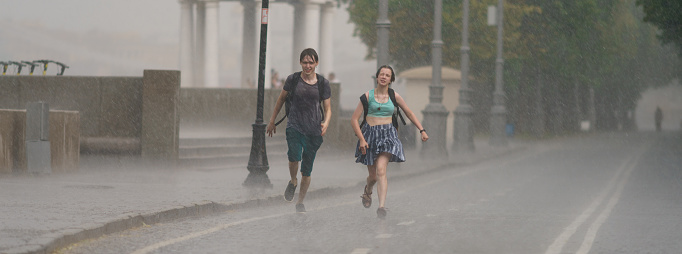Moscow, Russia. June 27, 2021: Raining day in city at summer. Wet young man and woman running. Texture of strong, fresh water drops and sprays. Tropical storm as a result of global warming. Panoramic