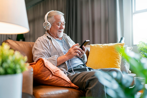 Asian Elderly man in headphones sitting on couch and listening to music singing along while resting in cozy living room at home,asian man using smartphone choosing playlist song from his application smartphone home casual lifestyle