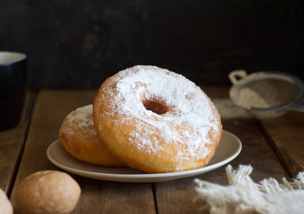 Two donuts with powder lie on a saucer on a wooden table. Nearby are a sieve, towel, walnut and mug. Rustic style