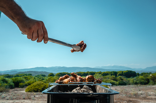 The background is green nature, the hills. The barbecue is in the main focus. With tongs, he raises the meat on the barbecue. The background is clean. Space for text.