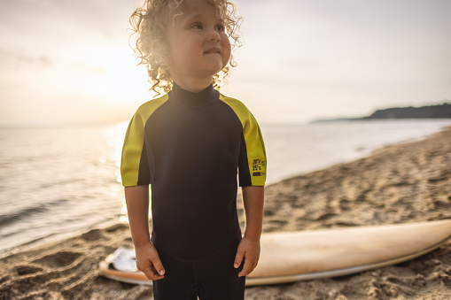 Portrait of little  blonde surfer boy standing on the beach in a wetsuit