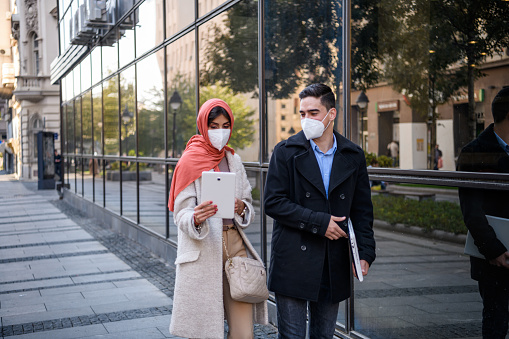 A mid adult Muslim businesswoman and a young Caucasian businessman are walking to work while wearing face masks.