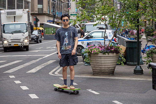 Midtown Manhattan, New York, NY, USA - July 5th 2022: Young man of Asian heritage with sunglasses driving down Broadway on a skateboard