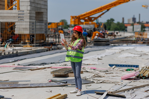 A young Caucasian female engineer wearing protective equipment is standing on a construction site and checking the plans on her tablet.