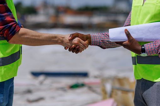 A close-up of a young Caucasian male engineer and a young African American male engineer shaking hands, while standing on a construction site.