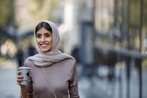 A mid adult Muslim businesswoman with a beautiful smile is walking down the street to work and carrying a cup of coffee.