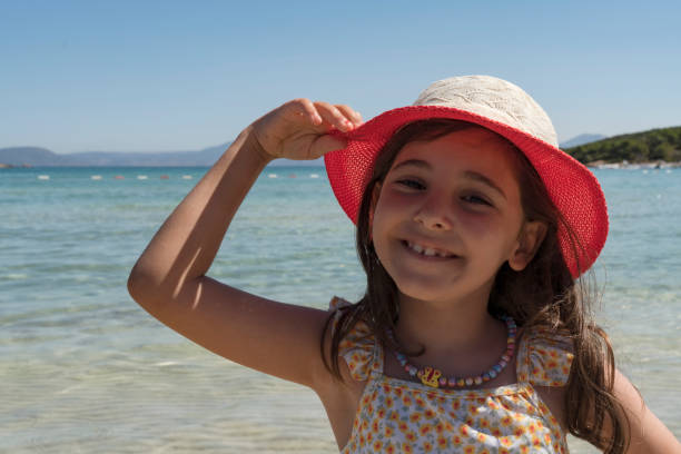 Young preschool child wearing a protective sun hat, standing on a sandy beach by the sea on a sunny summer day