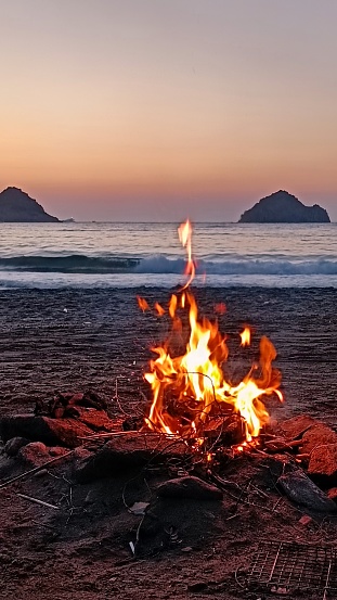 A vertical shot of a camp fire on the sandy beach by the Aegean sea. The time is right after sunset, the sky is colorful with red and earth tones. There are two small islands visible in the distance on the horizon. There is a metal grill partially visible in the front right corner.