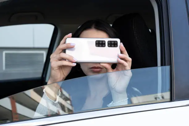 Photo of young woman takes pictures on the mobile phone from the car side window. a lot of cameras on smartphone. Spying with humor concept