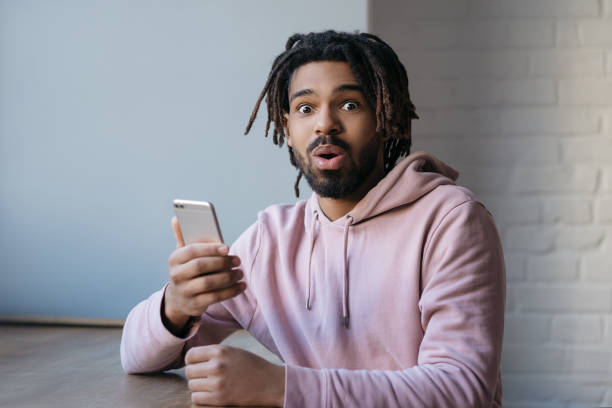 Surprised African American man holding smartphone, shopping online, booking tickets with sales stock photo
