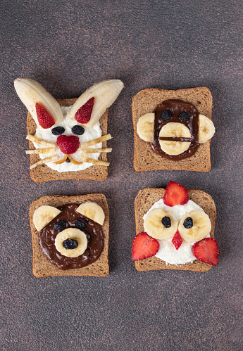 Four sweet toasts for kids in shape of chick, bear, rabbit and monkey, with strawberries, banana, cream cheese, chocolate and coconut flakes on brown background, Vertical format