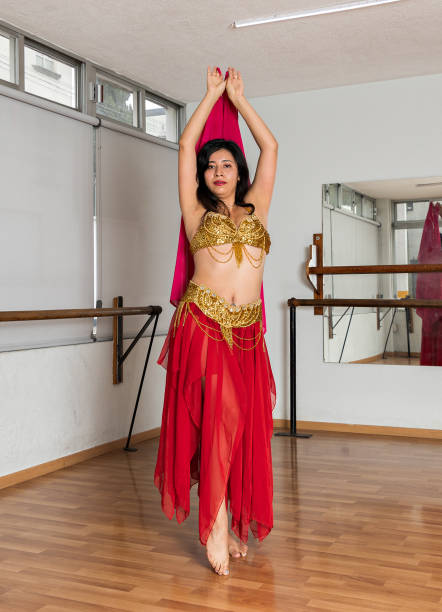 young bellydancer dressed in a red and gold costume practicing in a dance studio, twirling while moving a veil.