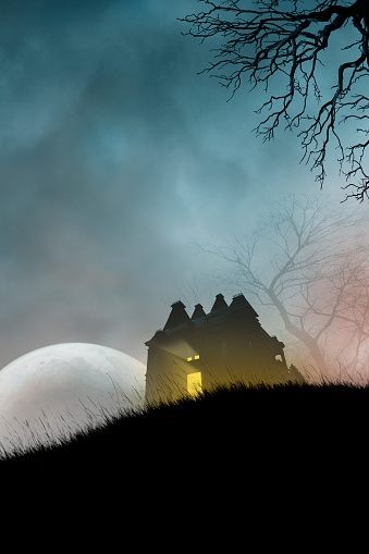 Light emanates from a haunted house on a grassy hill in front of a large waning moon that is framed by the twisted branches of an old bare tree on a dark and spooky Halloween night.