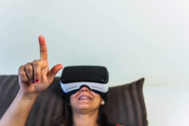 brunette woman with virtual reality goggles lying on bed holding up a finger to point at something. young girl looking at augmented reality through her device. - ai finger gamer imagens e fotografias de stock
