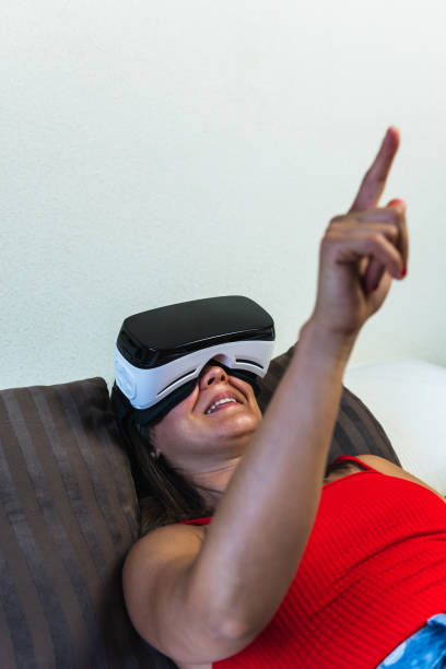 brunette woman with virtual reality goggles lying on bed holding up a finger to point at something. vertical image of young girl observing augmented reality through her device. - ai finger gamer imagens e fotografias de stock