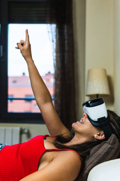 istock Woman lying in bed pointing at the ceiling with her virtual reality goggles on. Vertical image of young girl in her room trying out augmented reality while having a three dimensional experience. 1414672724