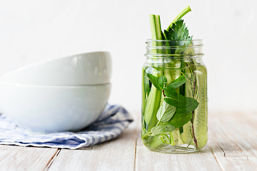 Infused water bottle with an abundance cucumber slices, mint leafs, celery stalk and rosemary in on white wood.