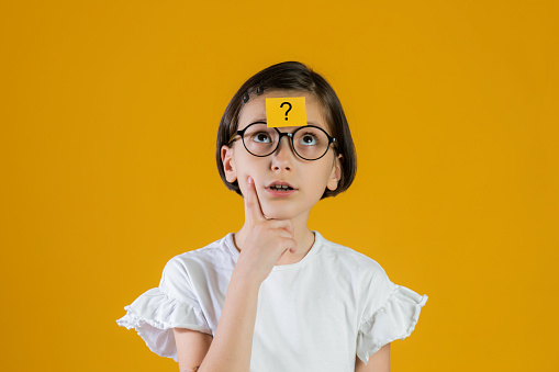 Little girl in white dress and eyeglasses looking up to a yellow note with question mark on her forehead in front of yellow background.