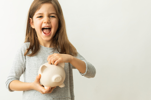 Little girl is laughing, holding piggy bank in hand and is looking at camera in front of white background.