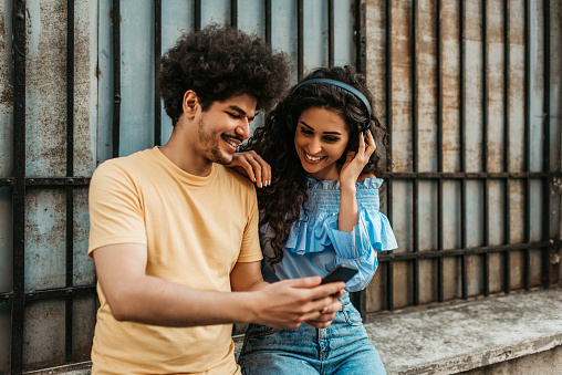 Young adult couple having good time together. They are walking down the street and laughing while listening to music with bluetooth headphones.