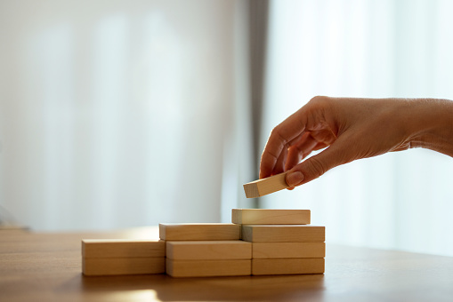 Close up photo of woman hand arranging wooden block stacking as step stair on a desk.