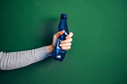 Close up photo of woman hand crushing a blue plastic bottle on a green background.