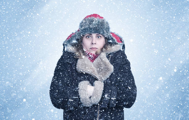 une jeune femme gelée en vêtements d’hiver se réchauffe les mains dans les manches d’un manteau en peau de mouton, il neige sur fond bleu - vêtement chaud photos et images de collection