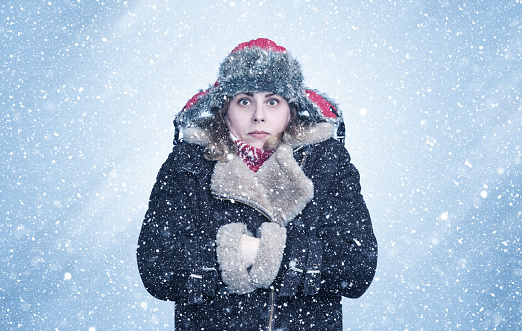 Frozen young woman in winter clothes warms her hands in the sleeves of a sheepskin coat, it's snowing around on a blue background