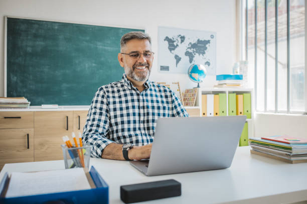 Elementary school teacher Teacher at classroom sitting at desk and using laptop. He is talking with students on video call. one mature man only audio stock pictures, royalty-free photos & images
