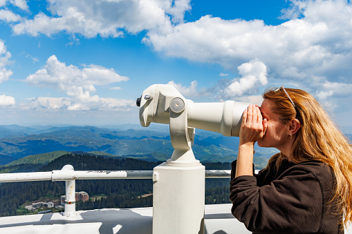 Curious girl tourist looks at mountainous wooded landscapes of mountain valley of Rhodope Mountains and blue cloudy sky through modern metal telescope, on high observation tower on top of Snezhan
