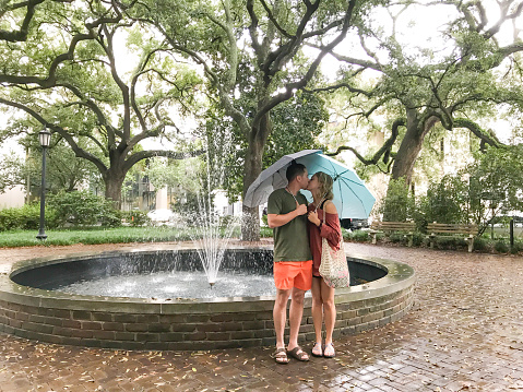 A young couple stands under an umbrella during a downpour in Savannah, Georgia. The couple is enjoying this vacation destination despite the rain.