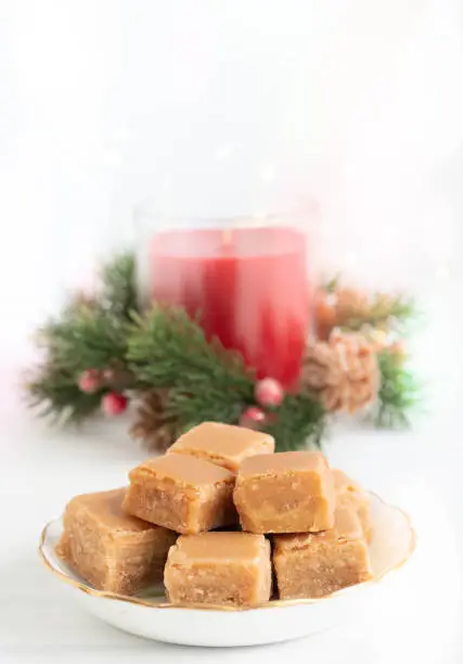 Caramel Fudge on Plate with Christmas Candle and Lights on Light Background with Copy Space Vertical