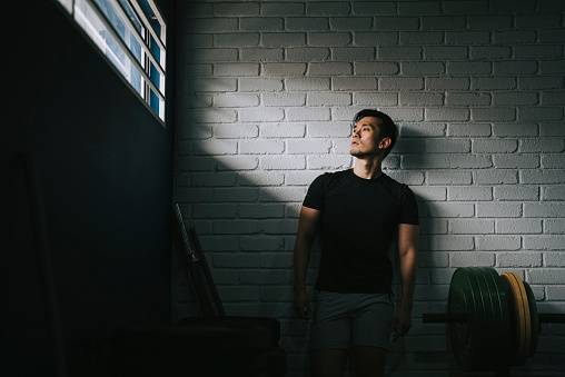 Asian Chinese Gay Man Resting leaning on brick wall in gym after routine workout looking away