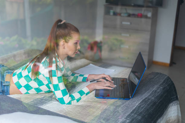 Beautiful Latin woman lying on her bed doing homework while taking her virtual class online