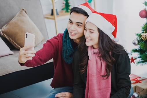 Young Asian couple lover wearing Santa Claus hat.Selfie with smartphone together in room decorated with Christmas trees at home background.Christmas celebration concept.