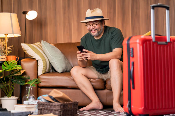 Ready to travel abroad,asia adult male man casual cloth sit on sofa couch next to luggage travel bag, hand reserve booking hotel room and flight ticket by online application smart device east travel listo para viajar al extranjero, asia hombre adulto hombre casual tela sentarse en el sofá sofá junto a la bolsa de viaje de equipaje, reservar a mano la habitación del hotel y el boleto de avión por aplicación en línea dispositivo inteligente east t