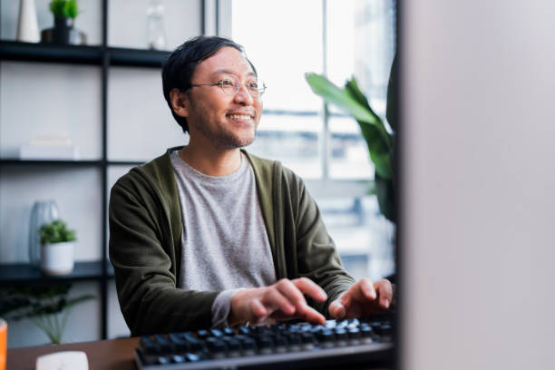 休日に自宅で働くアジアの成人男性、ホームオフィス:ハッピーヤングアジアのフリーランスのクリエイティブマン彼の家の快適さで働く、社会的距離の概念 - デスクトップ型パソコン ストックフォトと画像