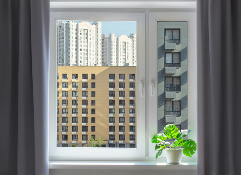 View through the window on facades of buildings. Dense urban development background. Interior space with dark grey curtains and indoor plant.