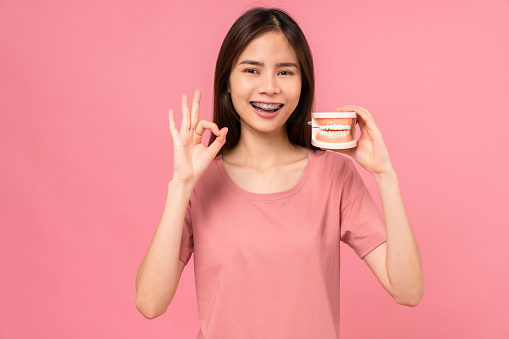 Mujer asiática sonriente que usa aparatos ortopédicos muestra un signo de ok y sostiene el modelo de dientes sobre fondo rosa, concepto de higiene oral y atención médica. photo