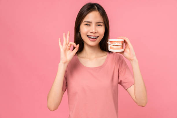 Smiling Asian woman wearing braces shows ok sign and holding tooth model on pink background, Concept oral hygiene and health care. stock photo