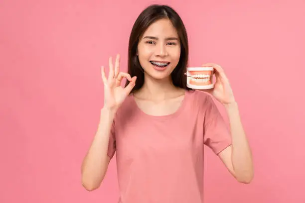 Smiling Asian woman wearing braces shows ok sign and holding tooth model on pink background, Concept oral hygiene and health care.