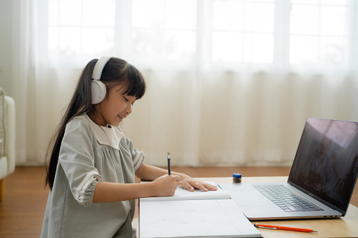 A young Asian girl studying at home  raises her hand to answer a teacher's question on a computer.