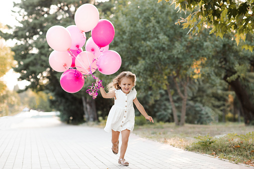Happy smiling child girl 3-4 year old wear casual white dress holding pink baloons run cheerful in park outdoor. Birthday celebration party. Childhood.