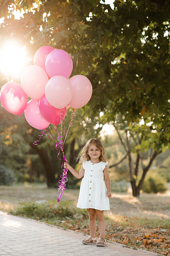 Happy smiling child girl 3-4 year old wear casual white dress holding pink balloons in park outdoor. Birthday celebration party. Childhood.
